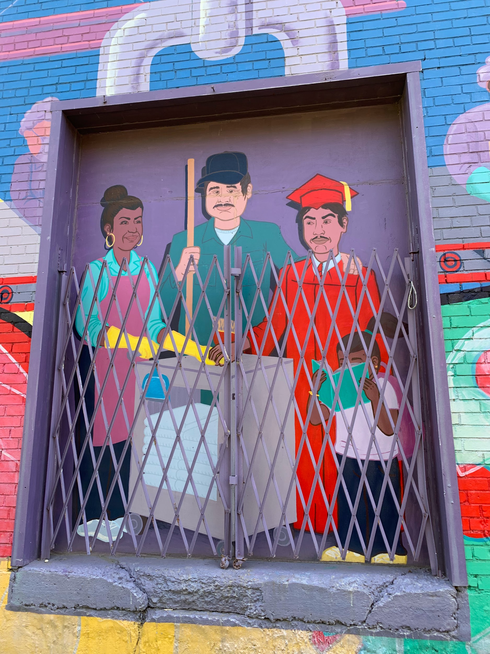 A zoomed in image of the family painted on the mural “El Abrazo.” The mother of the family holds cleaning supplies and is wearing a robe. The father is holding a broom. Their son is wearing a graduation cap and gown. Their daughter is reading a book.