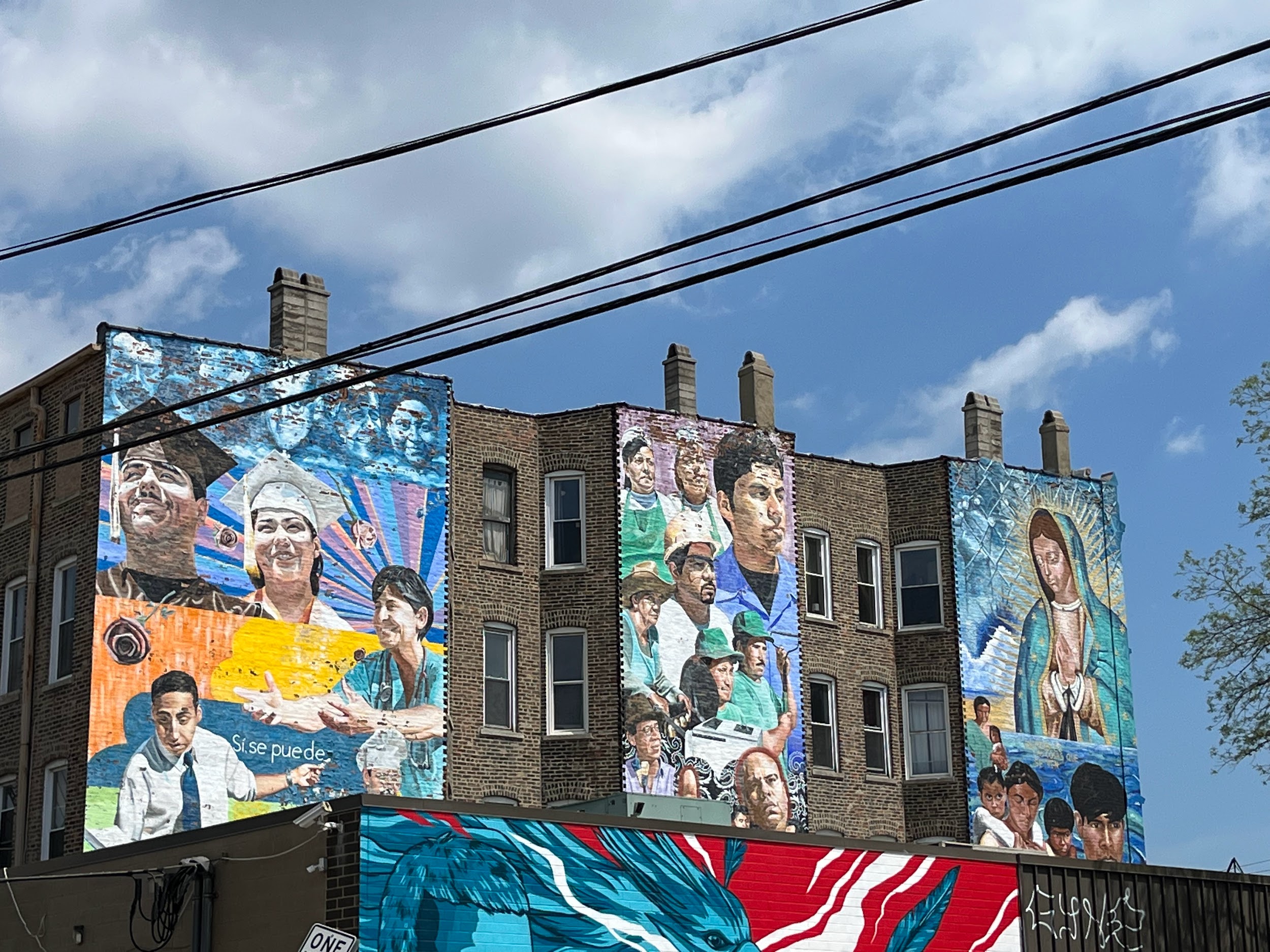 Alt text: This mural is “Las Cosas Q’Se Ven, three separate panels painted on the side of one building. On the first panel, two young graduates are smiling, with Latino historical figures above them. Below, a smiling doctor stretches out her hands, while a teacher chalks the words “Si, se puede” Yes, we can. A chef smiles below. In the middle panel, multiple people representing the working class community turned to the right, facing the last panel. The right-most panel depicts a large-scale painting of the Virgin of Guadalupe watching over immigrants sailing across the sea to the United States. Behind her, a man with his eyes closed grasps at a chainlink fence.