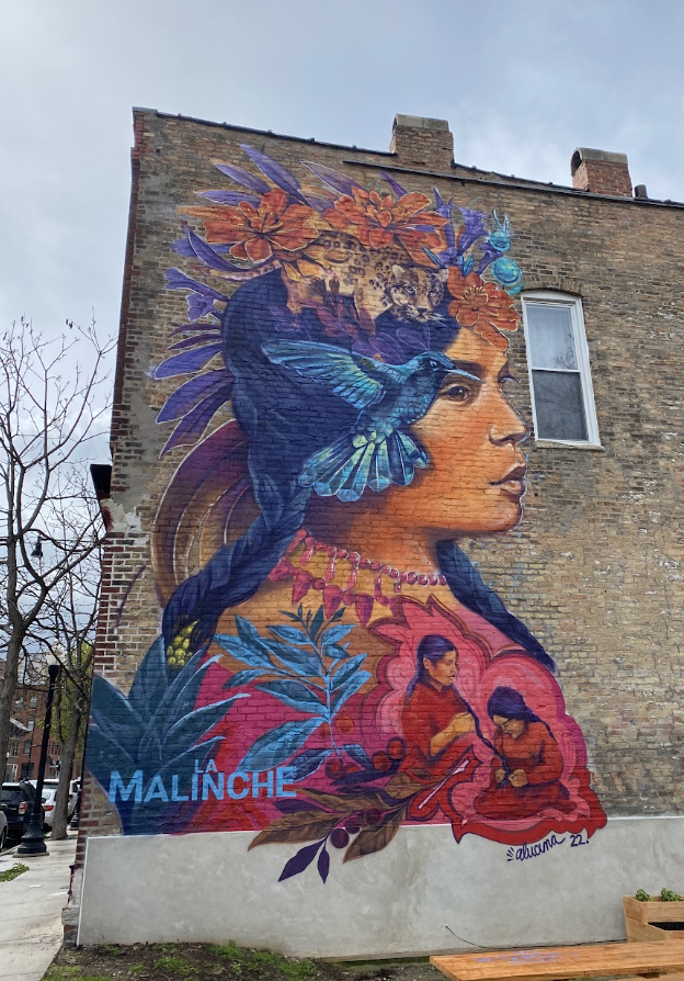 This mural is “La Malinche,” painted on the side of the cafe of the same name. It depicts a portrait of a woman who was the daughter of an Aztec Chief and later was sold to the conquistador Hernán Cortés as an interpreter. She has flowers in her hair, a hummingbird next to her face, and two women harvesting coffee beans below her.
