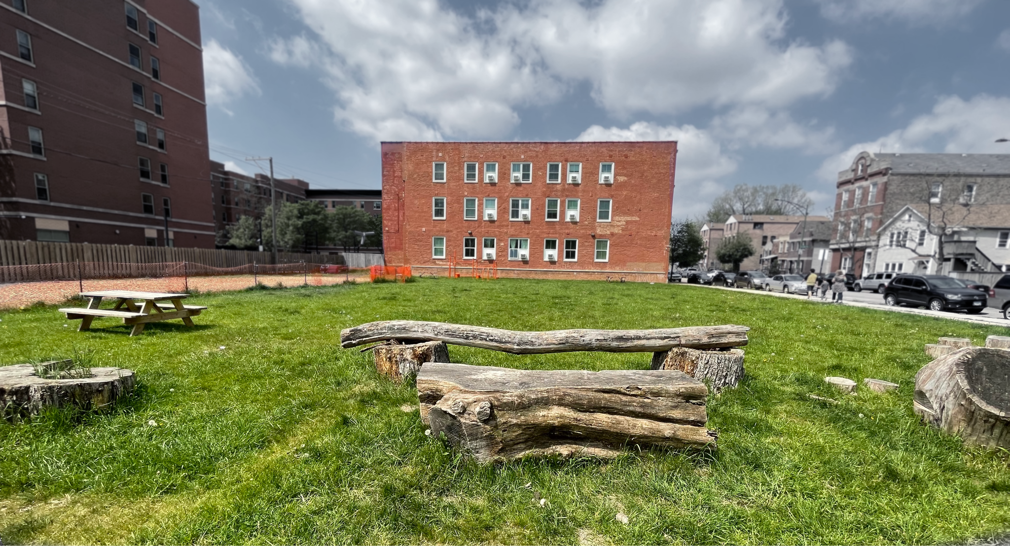 An image of a large, green, grassy yard with a short, orange three story building in the background. There is a picnic table to the left of the yard and tree stumps of various sizes scattered across the year close to the camera.