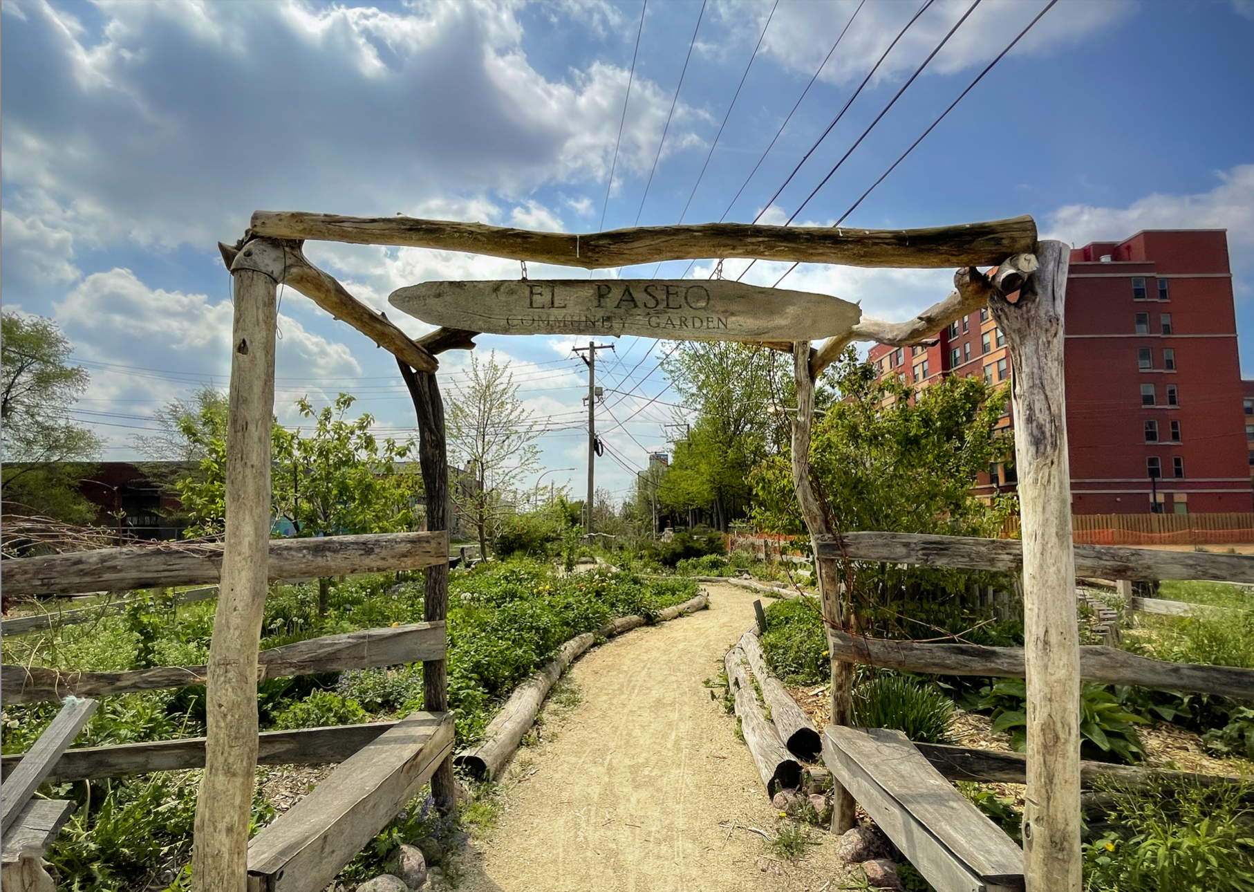 A picture of the permaculture site within the garden. A four poster wooden entryway with a sign reading “El Paseo Community Garden” in the middle. Lucious greenery is behind the entryway.